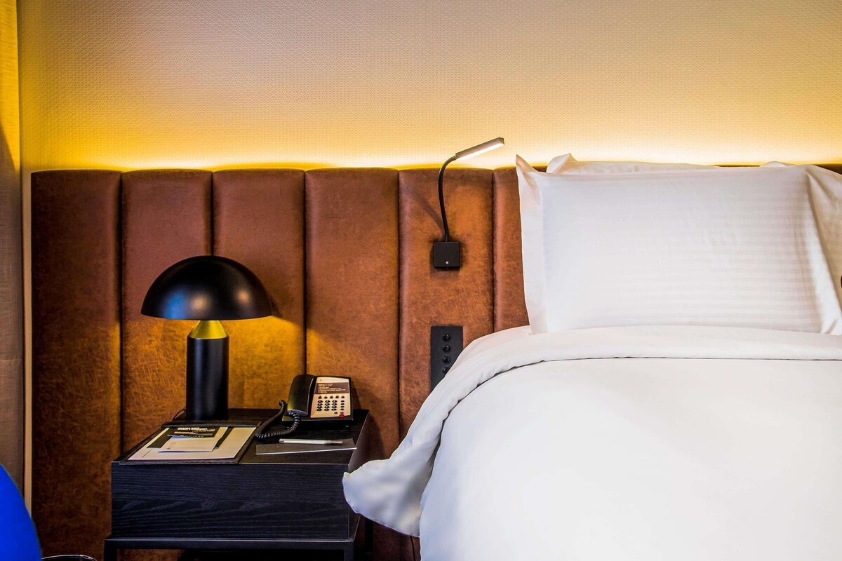 Een hotelkamer met een bed, een nachtkastje met een lamp, een telefoon en een wandcontactdoos voor opladen.