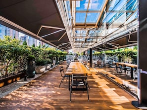 Een terras met een houten tafel en stoelen, omringd door potplanten en een glazen dakconstructie.