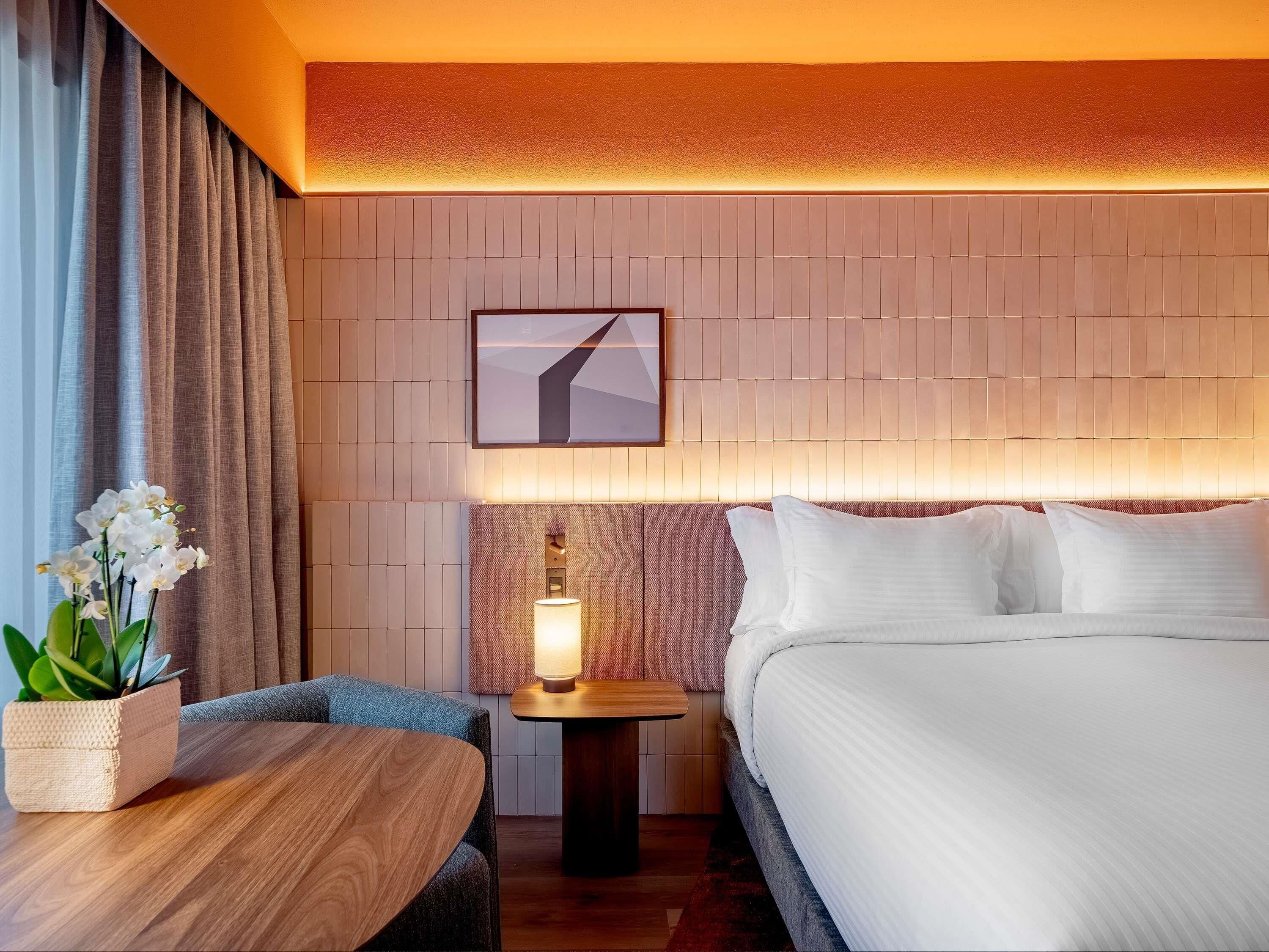 Een hotelkamer met een bed, een nachtkastje met een lamp, een klein tafeltje en een kamerplant.
