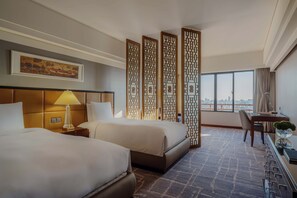 A hotel room with two beds, a desk, a chair, a lamp, a decorative screen, and a large window with a city view.