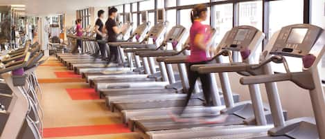 Un gimnasio con varias cintas de correr alineadas, ventanas amplias y suelo alfombrado.