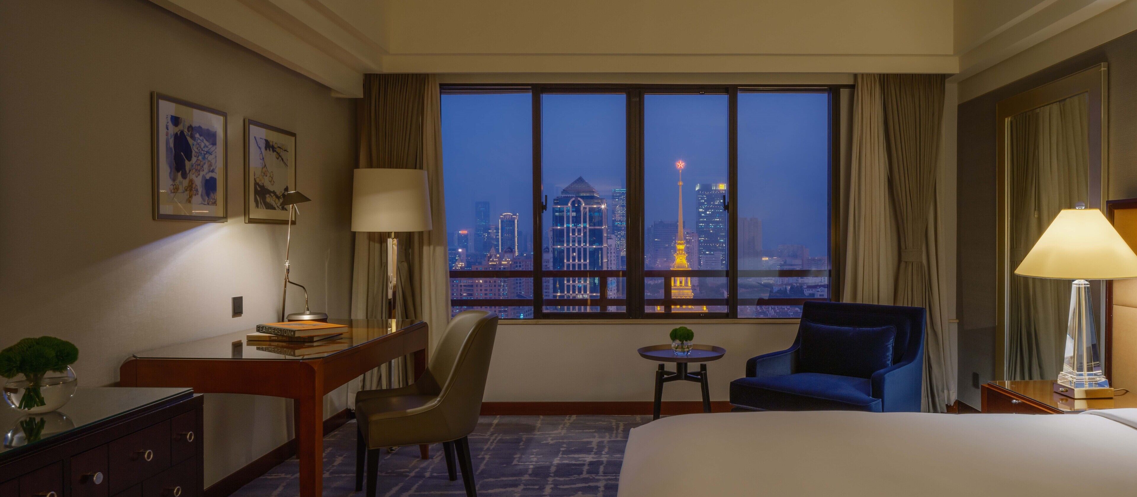 A hotel room with a large window offering a city view, a bed, a desk with a lamp, a chair, and a small table with a plant.