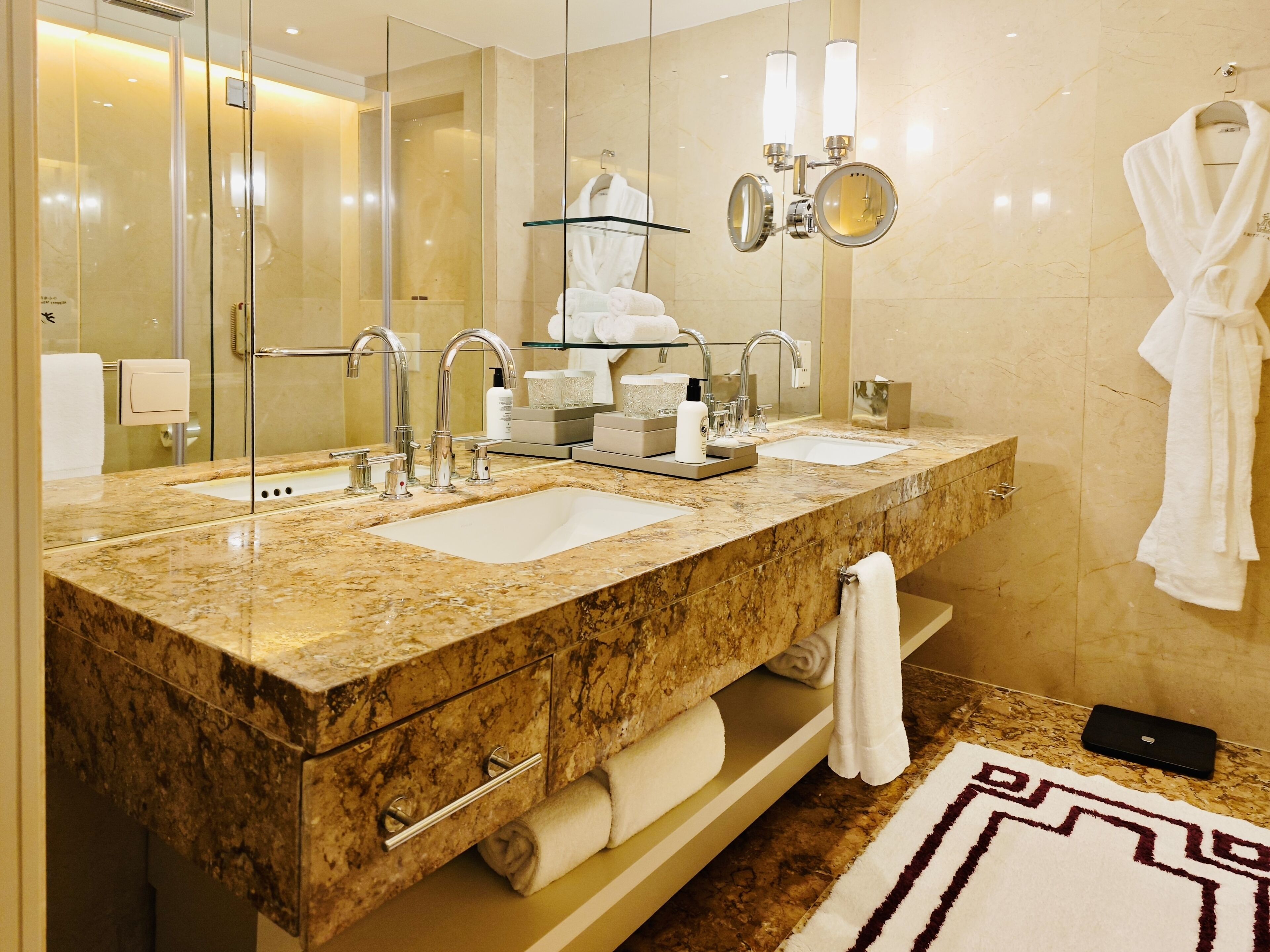 A modern bathroom with two sinks, a glass shower enclosure, a towel rack, and a mirror with lights.