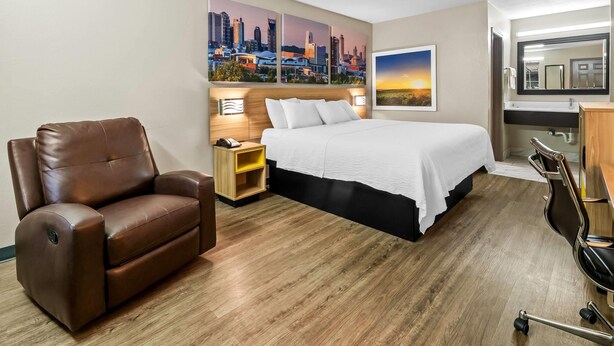 A hotel room with a bed, a brown leather chair, a nightstand, a desk with a chair, and a cityscape painting on the wall. - Days Inn by Wyndham Simpsonville (Simpsonville)