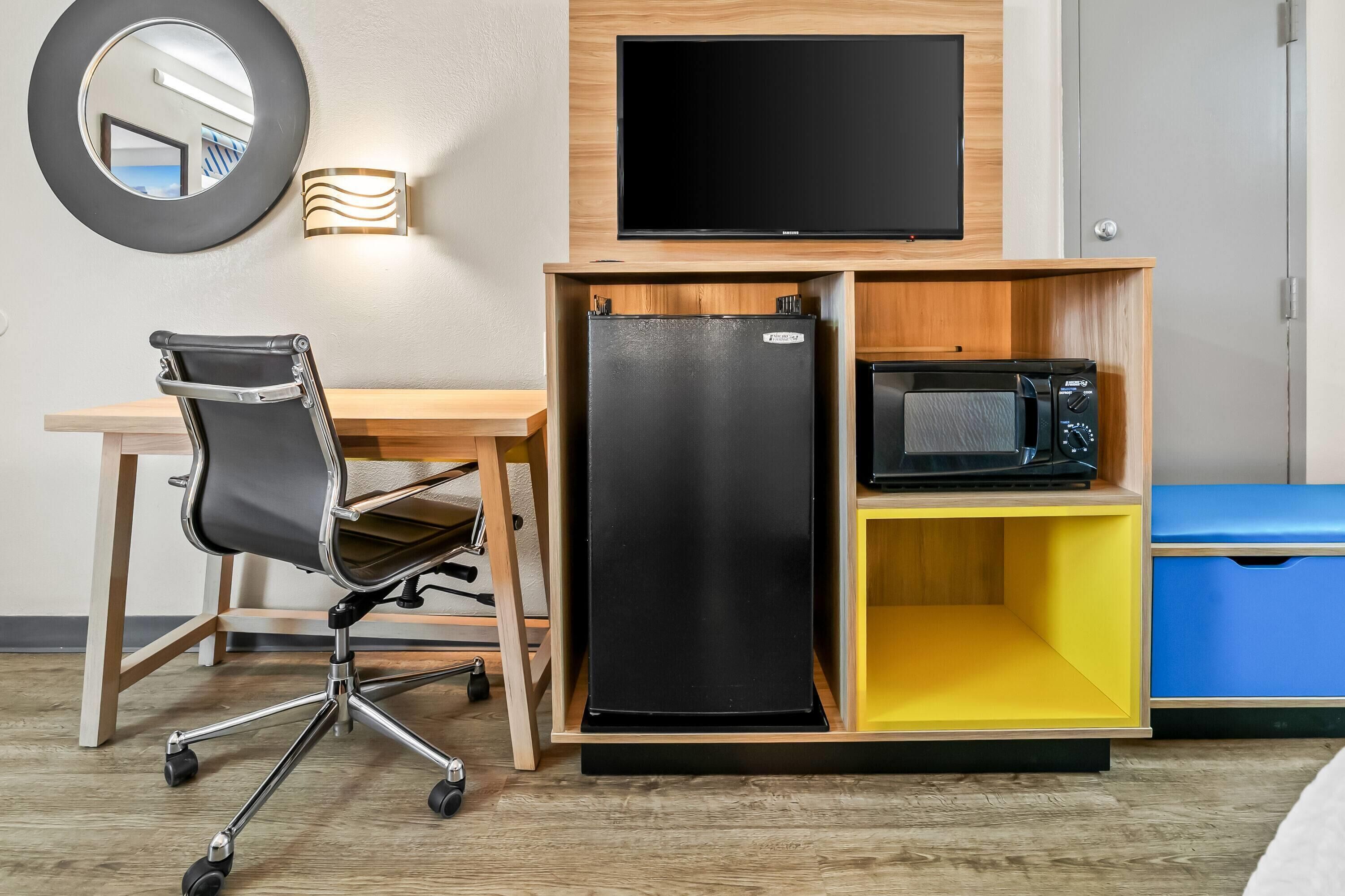 A compact room with a desk, chair, flat-screen TV, refrigerator, microwave, and a small storage unit.
