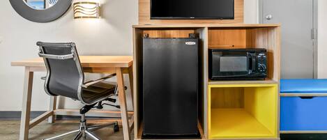 A compact room with a desk, chair, flat-screen TV, refrigerator, microwave, and a small storage unit.
