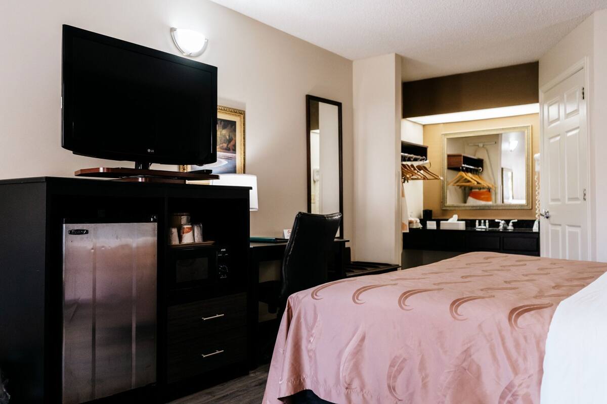 Habitación de hotel con cama, escritorio con computadora, televisor, espejo, armario y refrigerador pequeño.