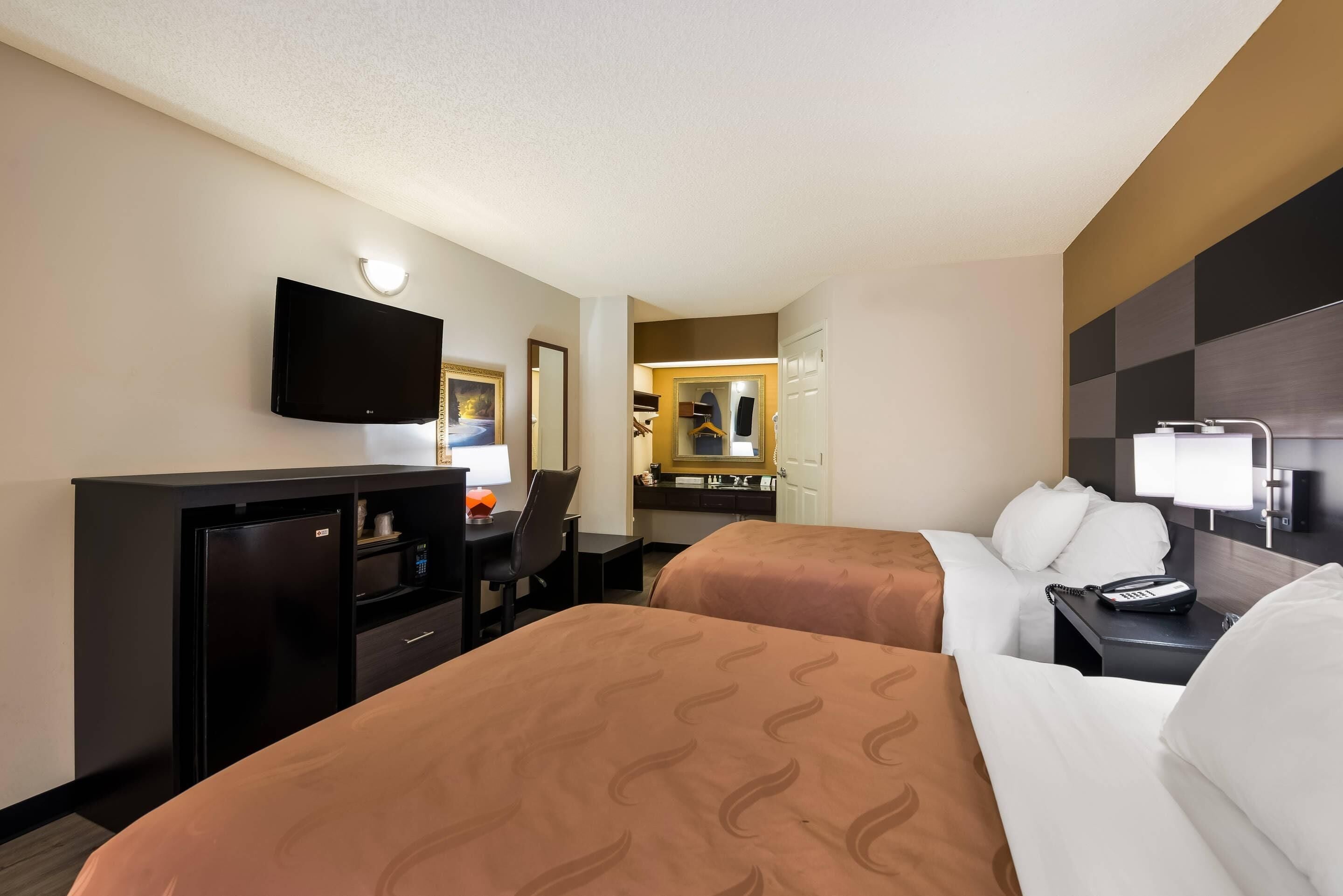 A hotel room with two beds, a desk with a computer, a TV mounted on the wall, and a mirror.