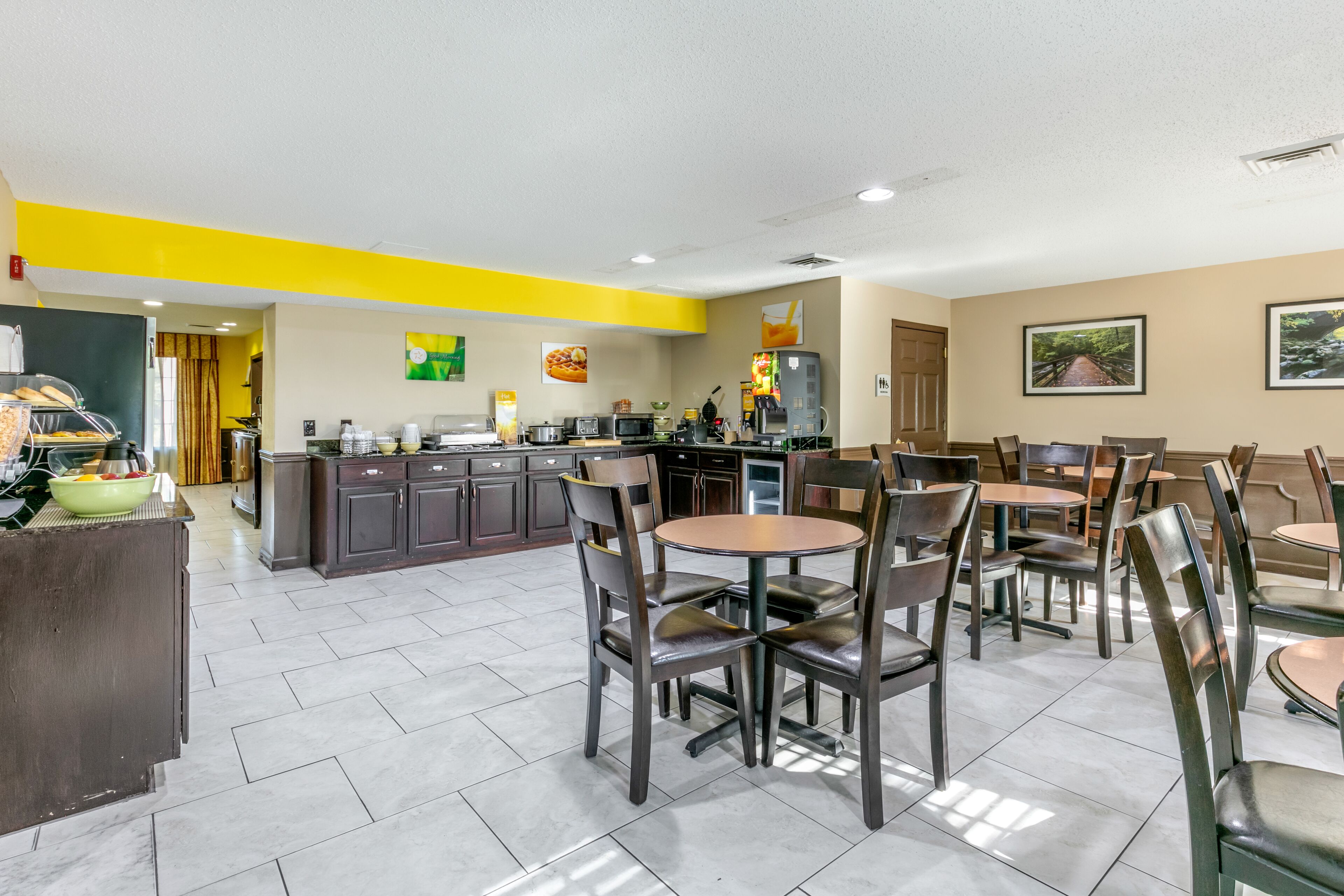 A modern restaurant with a bright yellow accent wall, a kitchen area, and several tables and chairs.