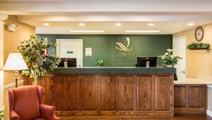 A reception desk with a wooden panel design, a green wall with the 'Quality Inn' logo, and a reception area with a chair and a plant.