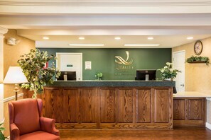 A reception desk with a wooden panel design, a green wall with the 'Quality Inn' logo, and a reception area with a chair and a plant.
