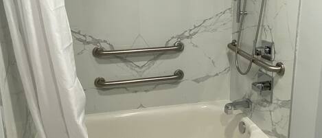 A modern bathroom with a marble wall, a bathtub, a shower with a handheld showerhead, and grab bars for accessibility.