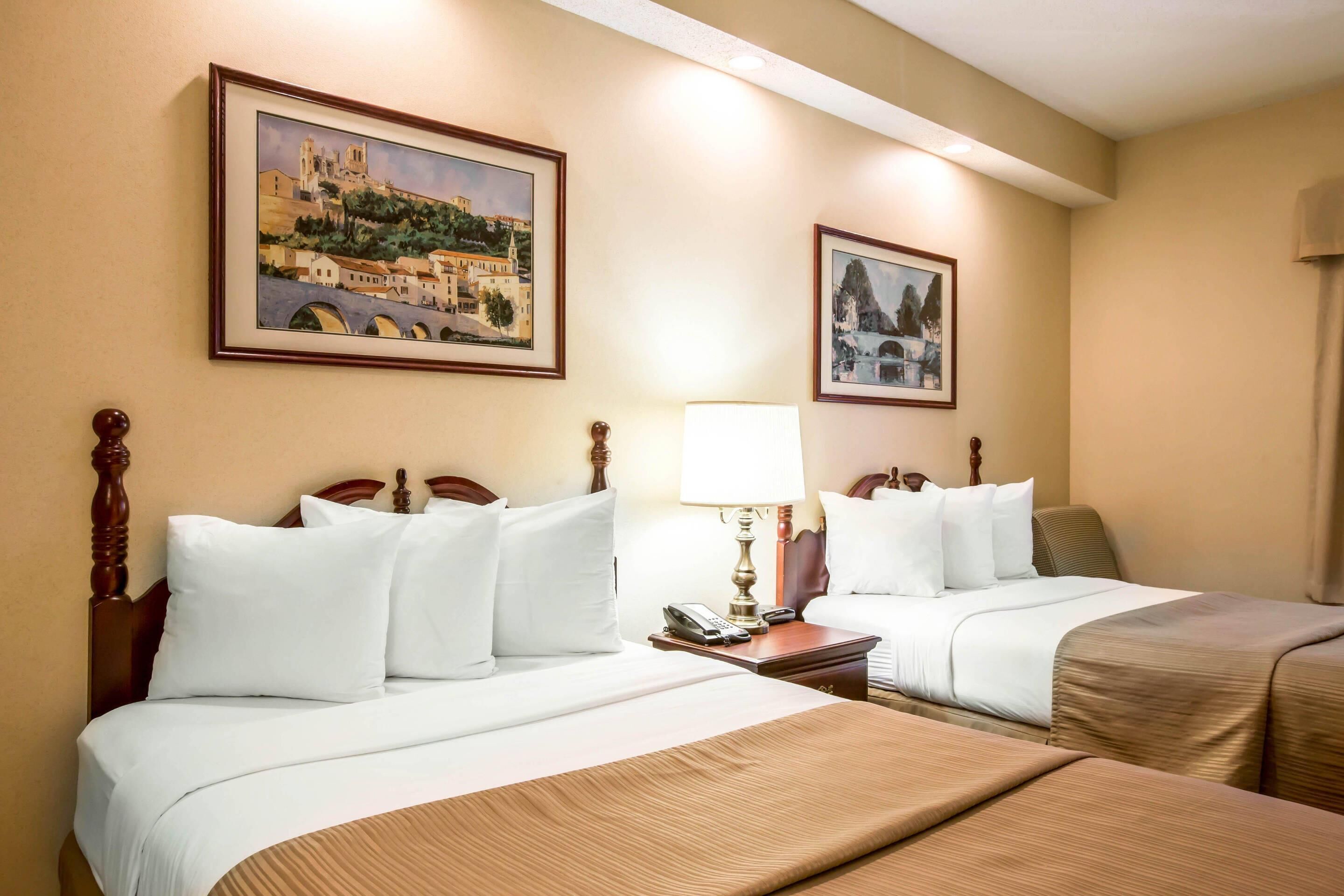 A hotel room with two beds, a nightstand with a lamp, and two framed paintings on the wall.