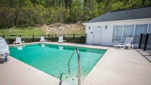 A rectangular swimming pool with clear water, surrounded by a concrete deck. There are lounge chairs and a covered area with a table and chairs. A metal ladder is in the pool.
