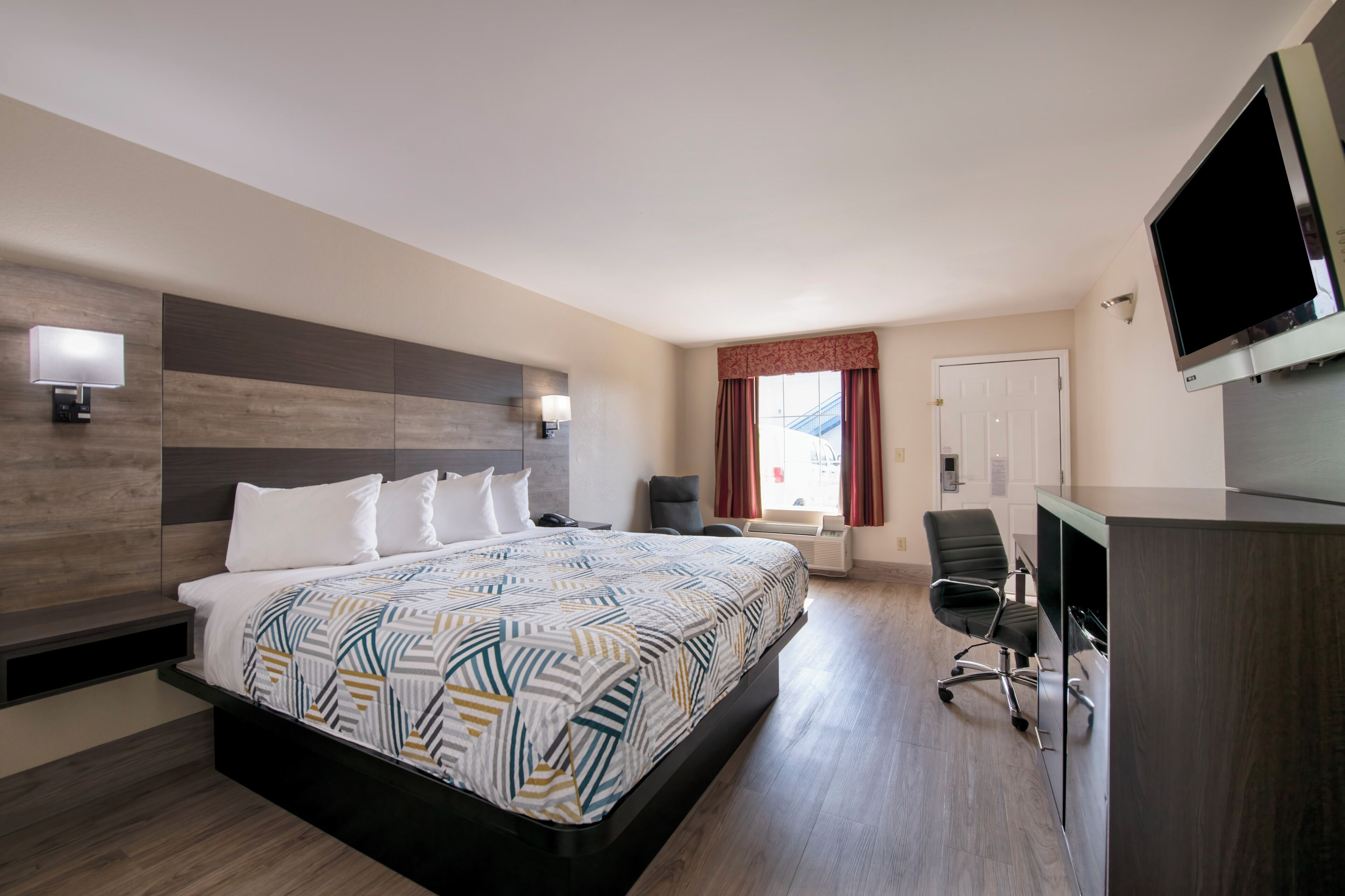 A hotel room with a large bed, a desk, a chair, a TV, and a window with curtains.