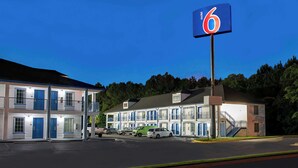 Un motel reconnaissable à son grand panneau indiquant le chiffre « 6 », avec de nombreuses chambres aux portes bleues et un parking où sont stationnés plusieurs véhicules.