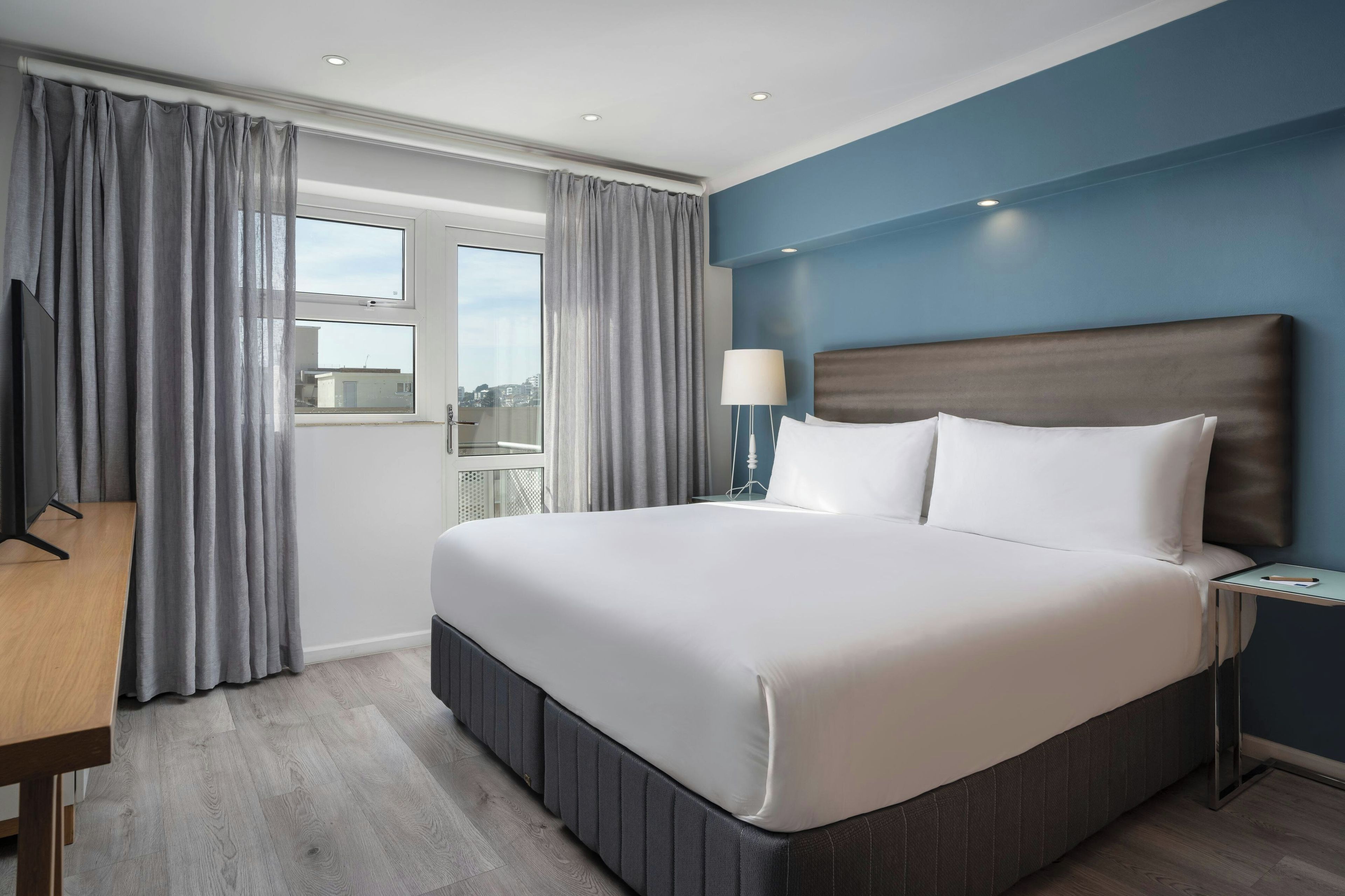 A hotel room with a large bed, a desk, a nightstand, and a window with curtains.