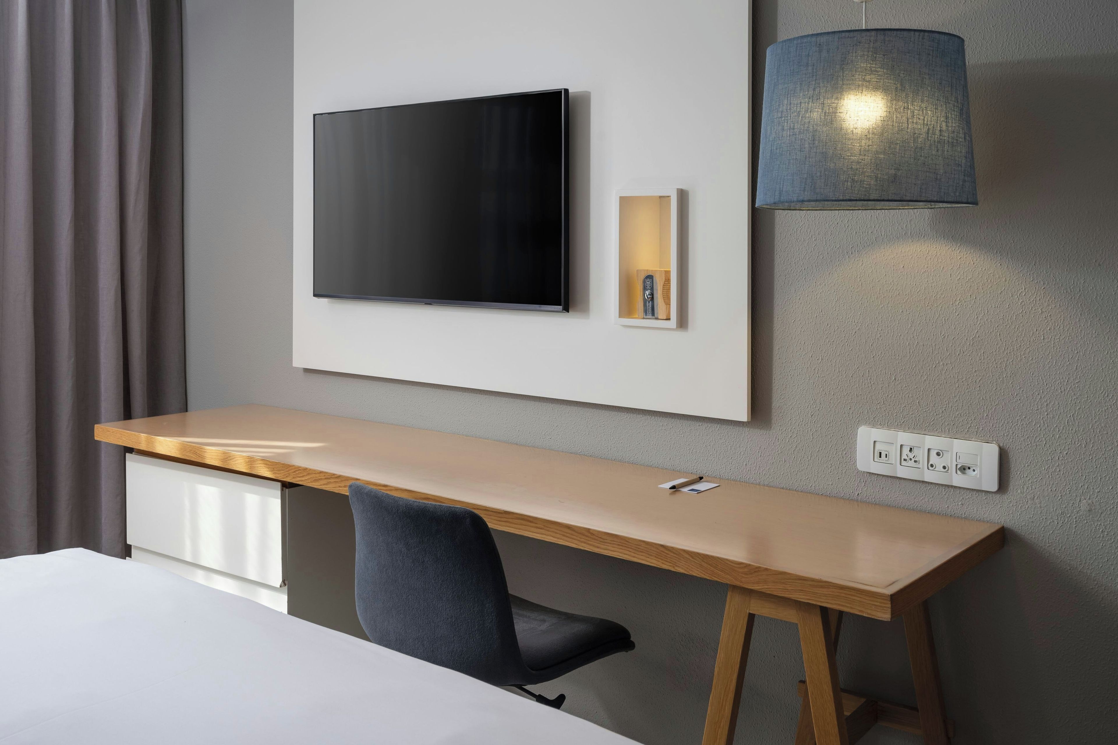 A modern hotel room with a wooden desk, a flat-screen TV mounted on the wall, a small bedside table with a lamp, and a power outlet.