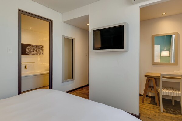 Una habitación de hotel con cama, televisión montada en la pared, una silla de madera y un espejo.