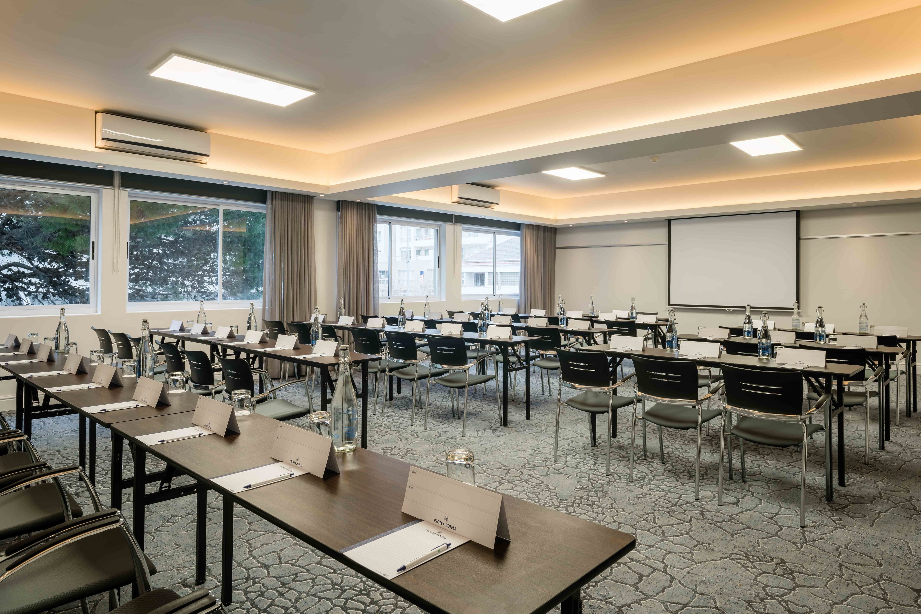 A conference room with multiple tables and chairs, large windows, and a projector screen.