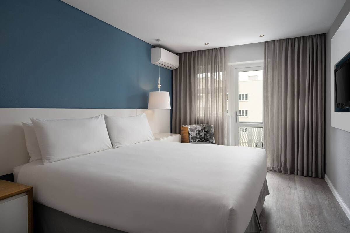 A hotel room with a large bed, a nightstand, a lamp, a TV, and a view of the outside through a window with curtains.