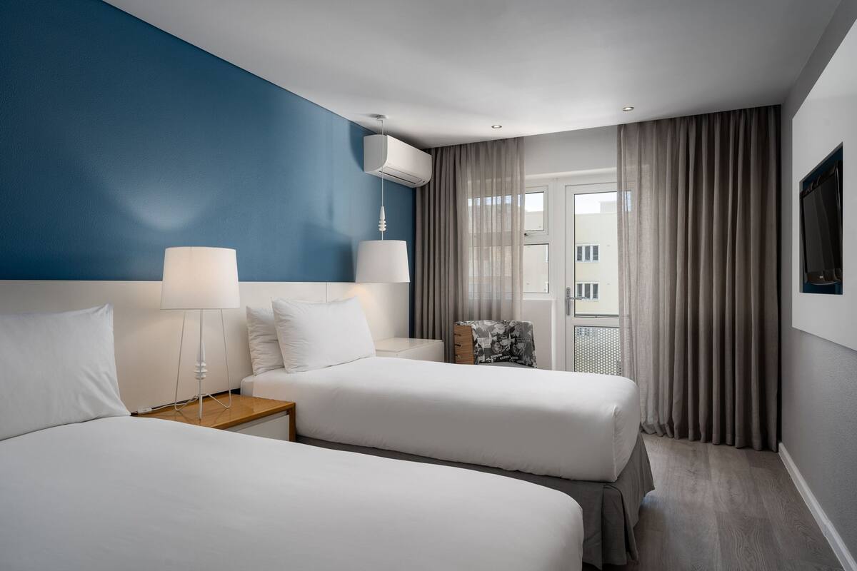 A hotel room with two beds, a nightstand, a lamp, a TV, and a window with curtains.