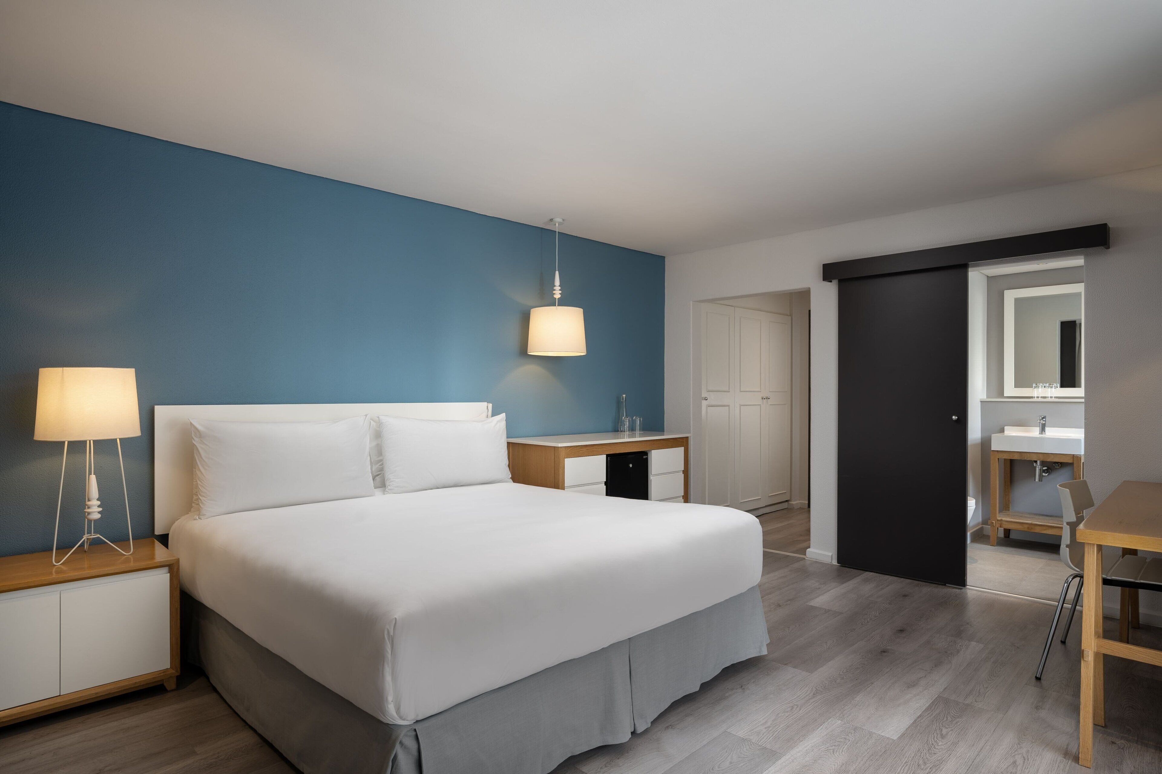 A modern hotel room with a large bed, bedside tables with lamps, a desk, a bathroom with a mirror, and a wardrobe.
