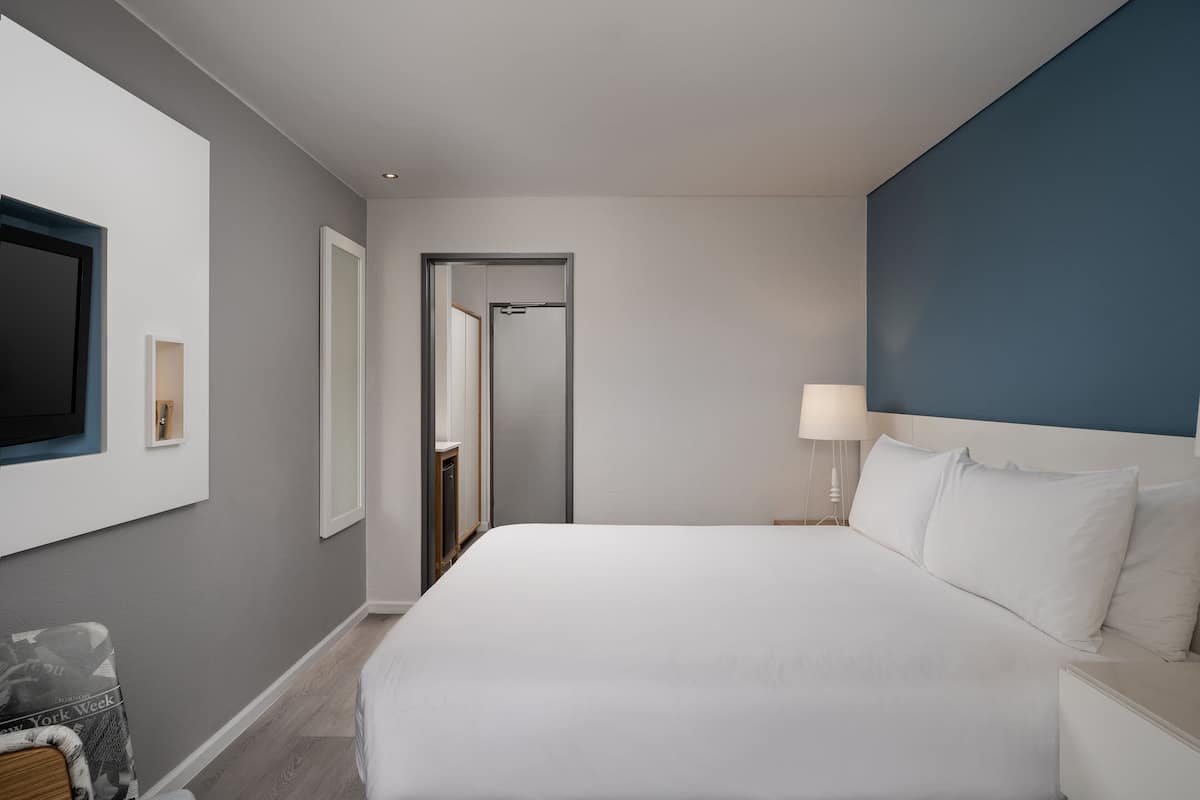 A hotel room with a large bed, a TV mounted on the wall, a bedside table with a lamp, and a door leading to a bathroom.
