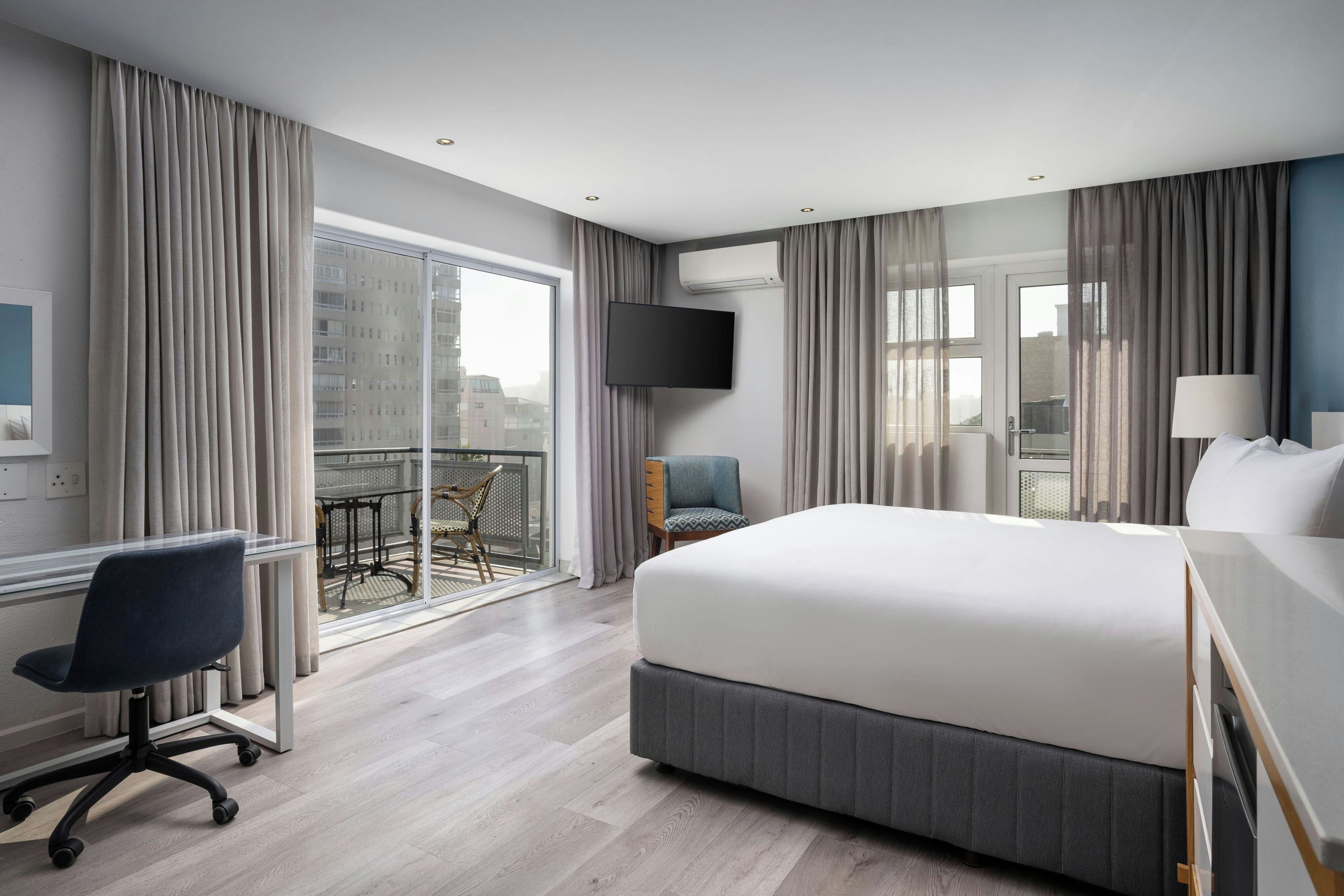 A hotel room with a large bed, a desk, a chair, a TV, and a balcony with a view of buildings.