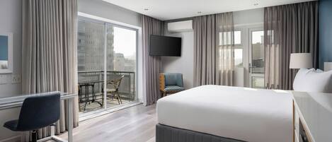 A hotel room with a large bed, a desk, a chair, a TV, and a balcony with a view of buildings.