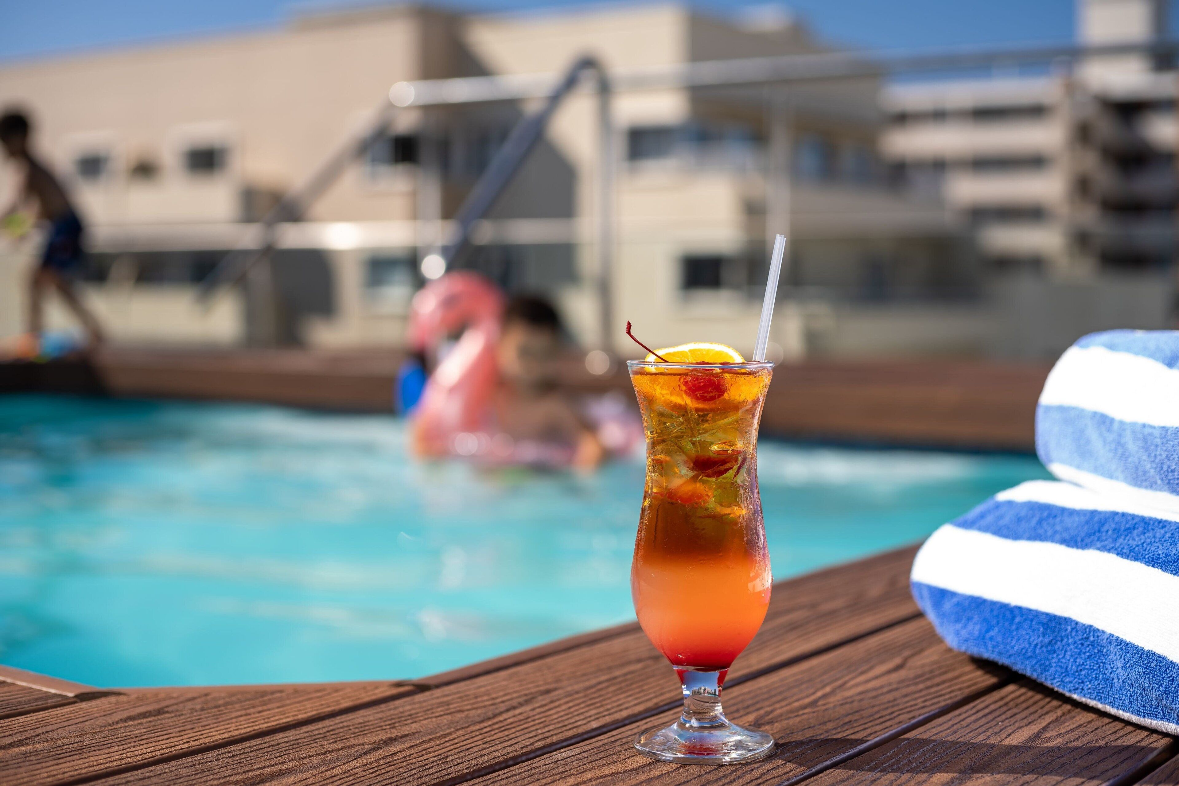 A cocktail glass with a straw and a slice of orange on a wooden deck by a pool.