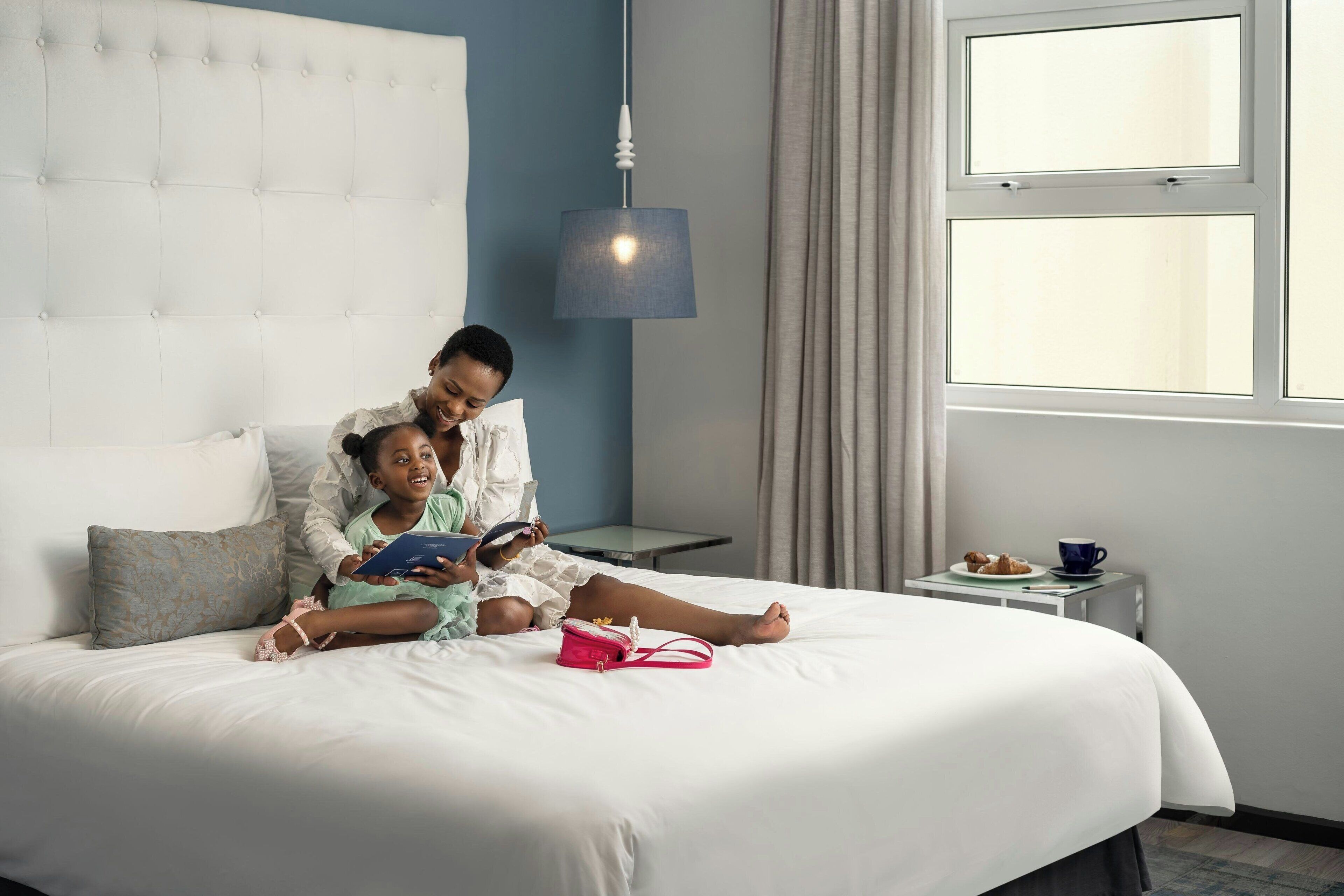 A hotel room with a large bed, a bedside table with a lamp, a window with curtains, and a small table with a cup and pastries.