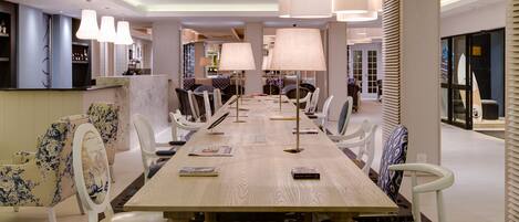 A modern hotel lobby with a long dining table, white chairs, and a large lamp.