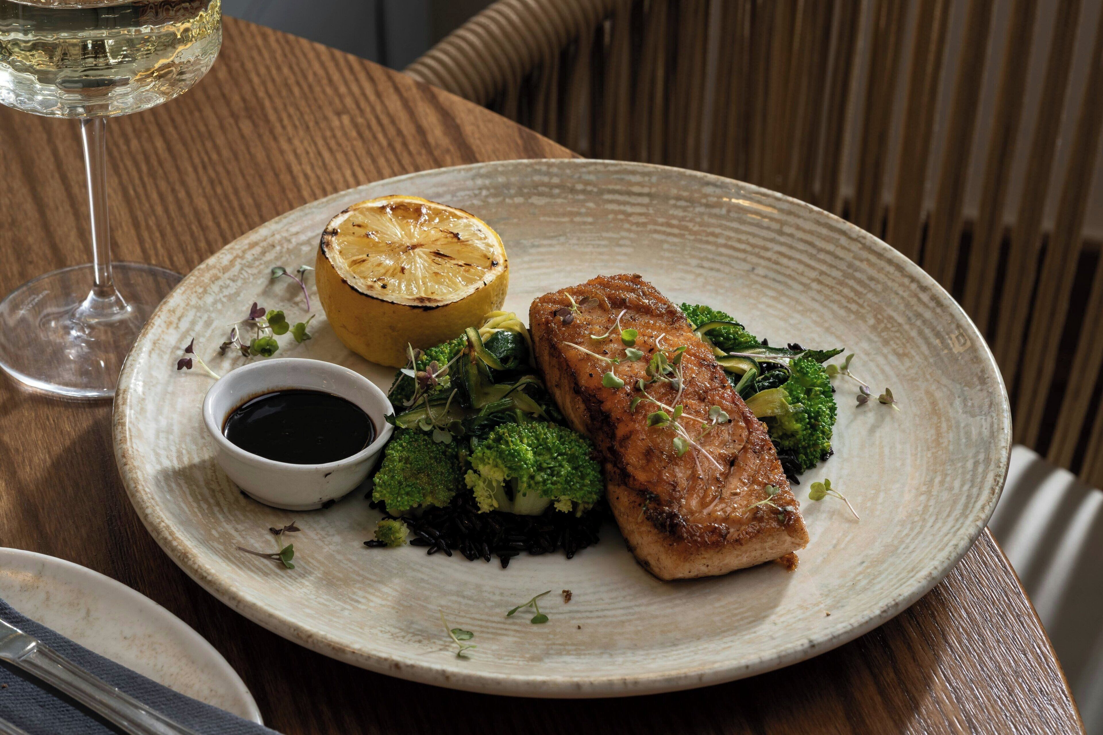 Een bord met gegrilde zalm, broccoli en een halve citroen, vergezeld van een glas witte wijn en een klein schaaltje saus, op een houten tafel.