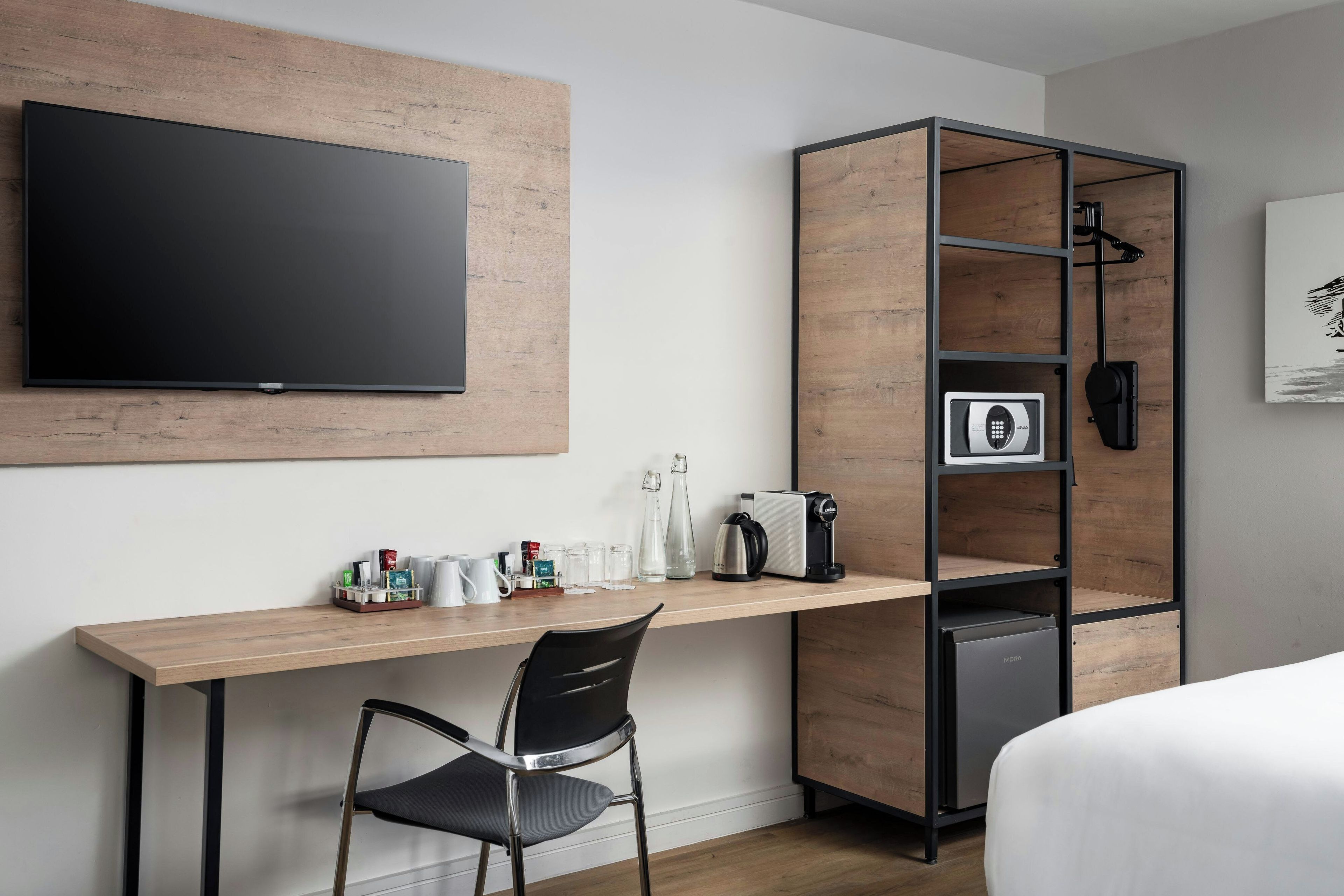 A hotel room with a wooden desk, a black chair, a flat-screen TV mounted on the wall, a minibar, and a bed with white bedding.
