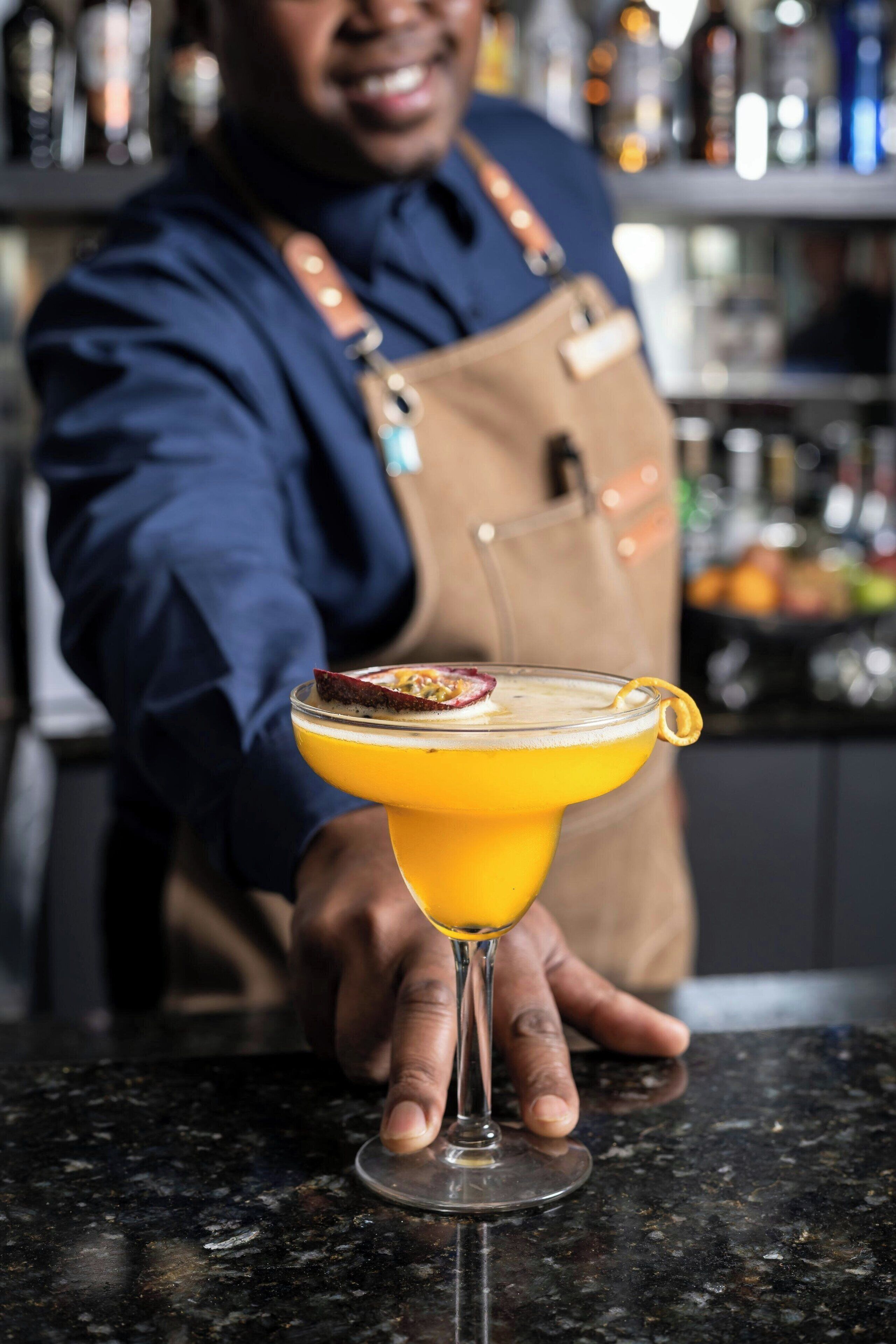 A bartender holding a cocktail with a garnish, in a bar setting with shelves of bottles in the background.