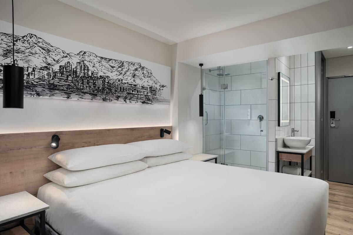 A modern bedroom with a large bed, a cityscape mural, a wooden headboard, a bedside table, a glass shower enclosure, and a bathroom with a sink and mirror.