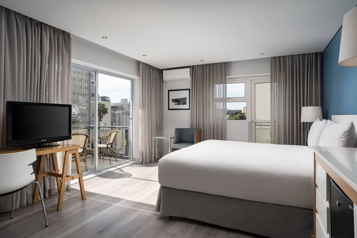 A modern hotel room with a large bed, a TV, a desk, and a balcony with a city view.