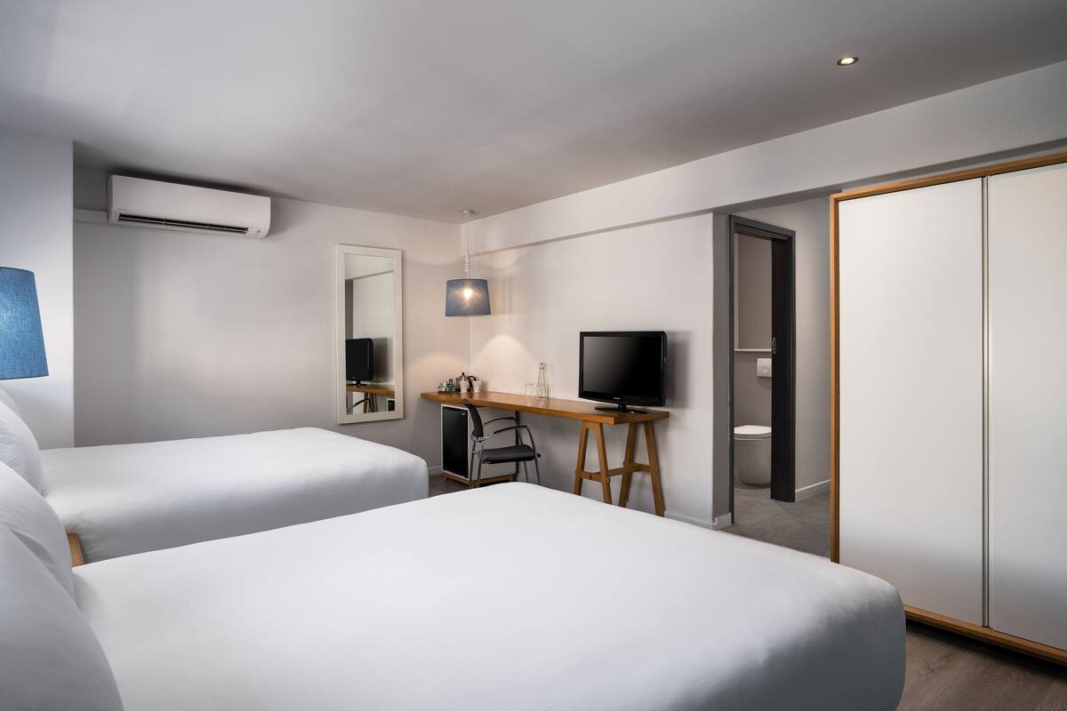 A hotel room with two beds, a desk with a television, a mirror, and an air conditioning unit.