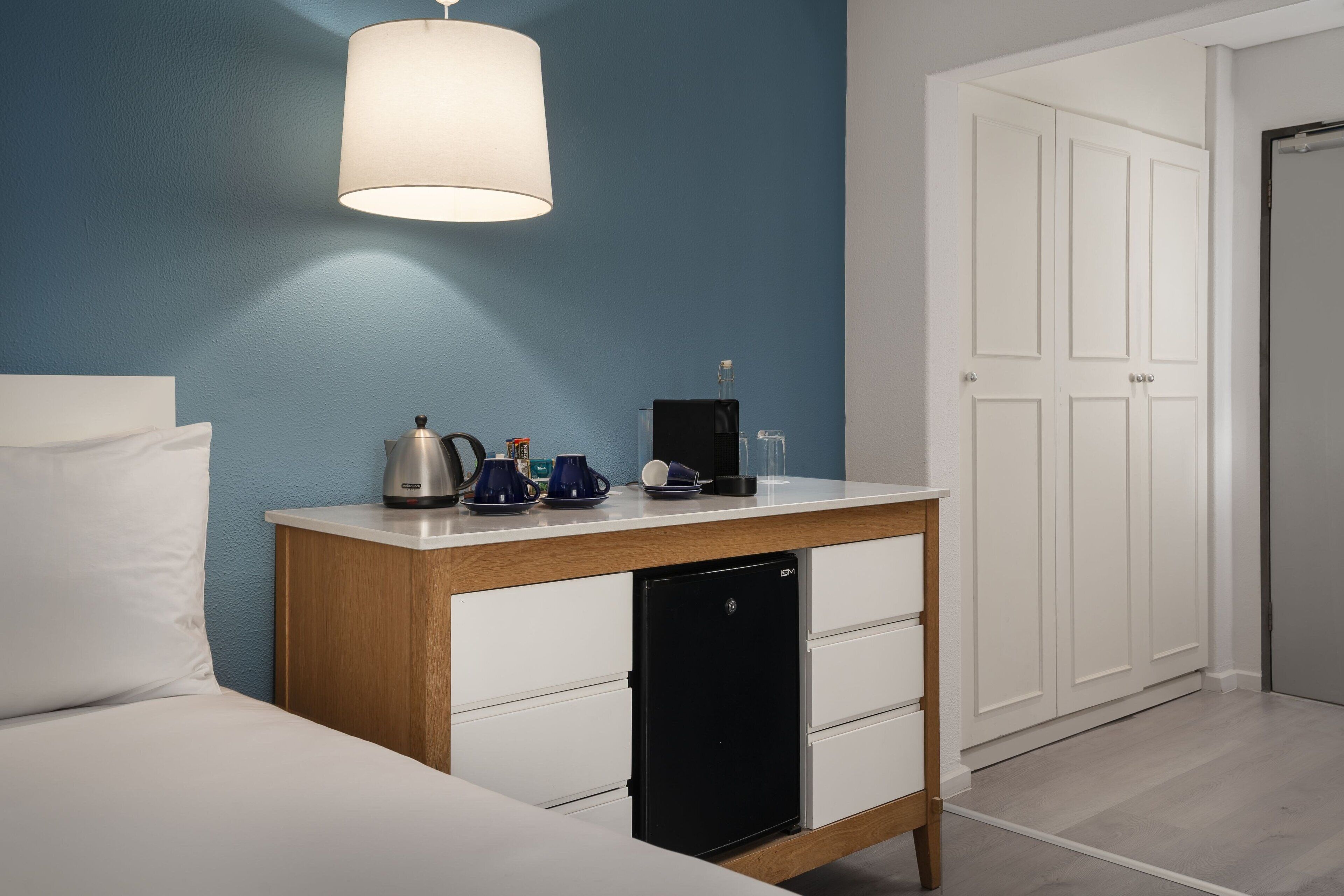 A bedroom with a bed, a small table with a lamp, a refrigerator, and a white wardrobe.