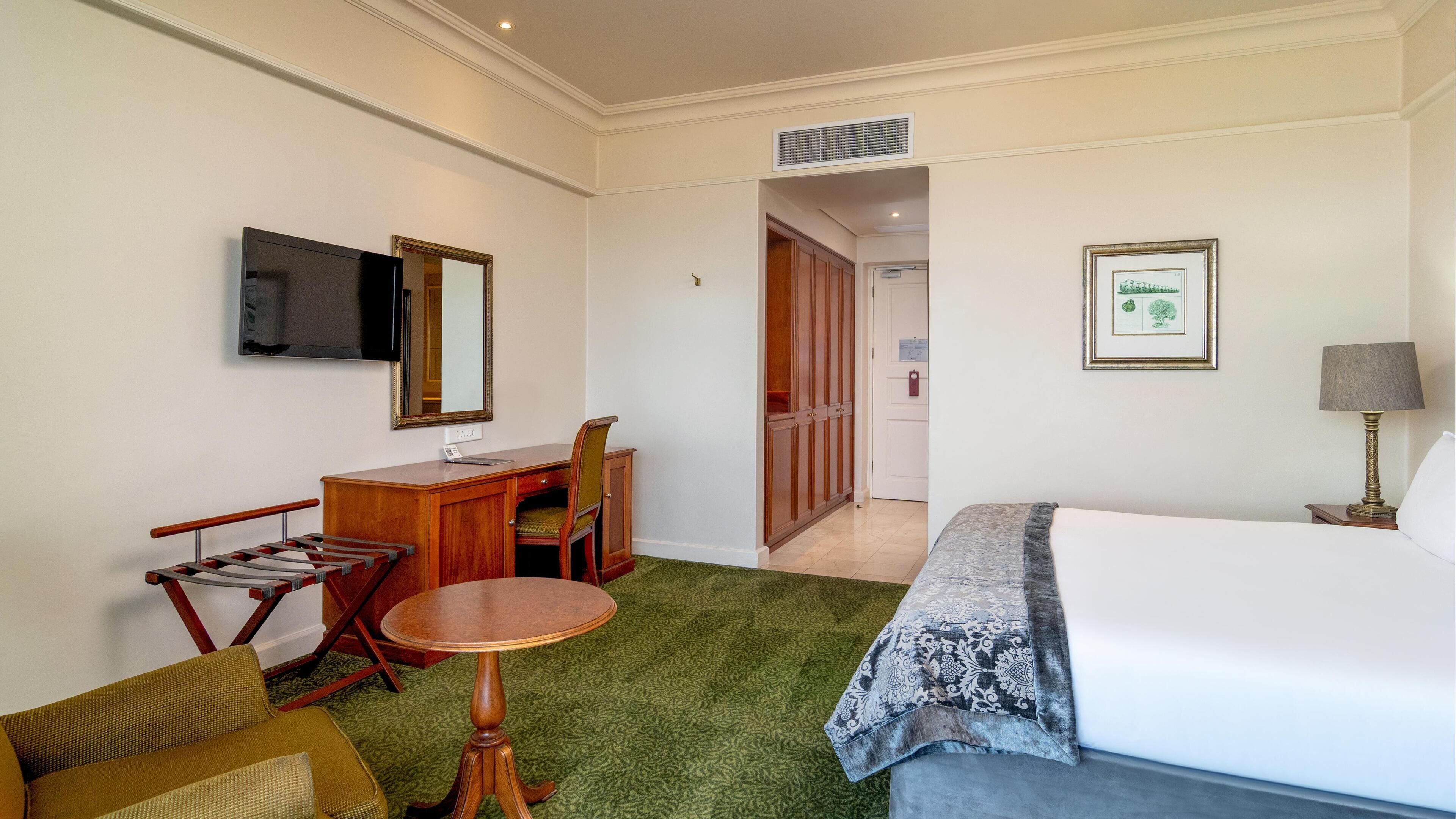 Une chambre d’hôtel comprenant un lit, un bureau, une chaise, une télévision, un miroir, un tableau encadré et une lampe.
