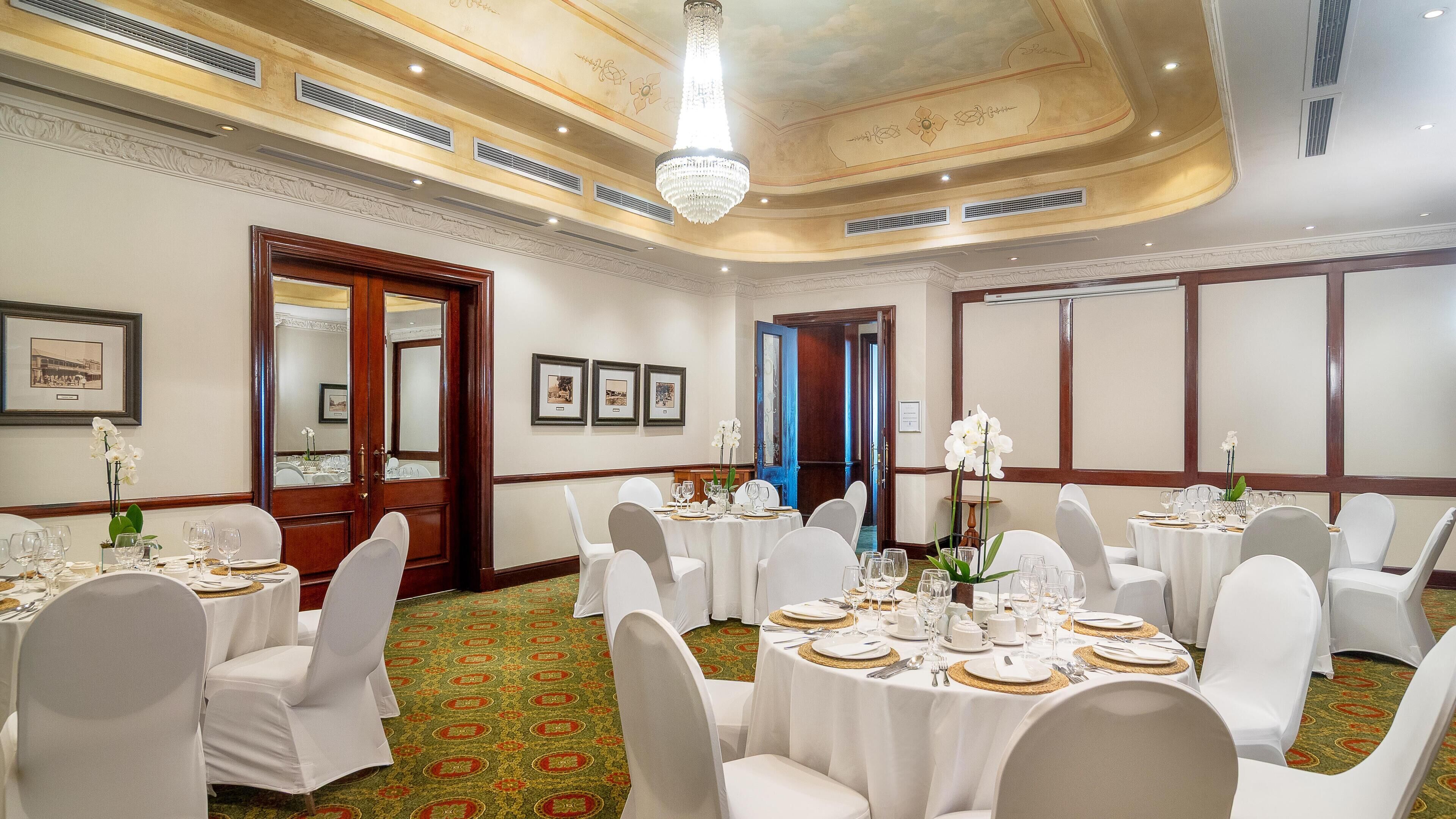 Un salón de banquetes con mesas redondas, sillas blancas y una araña de cristal.
