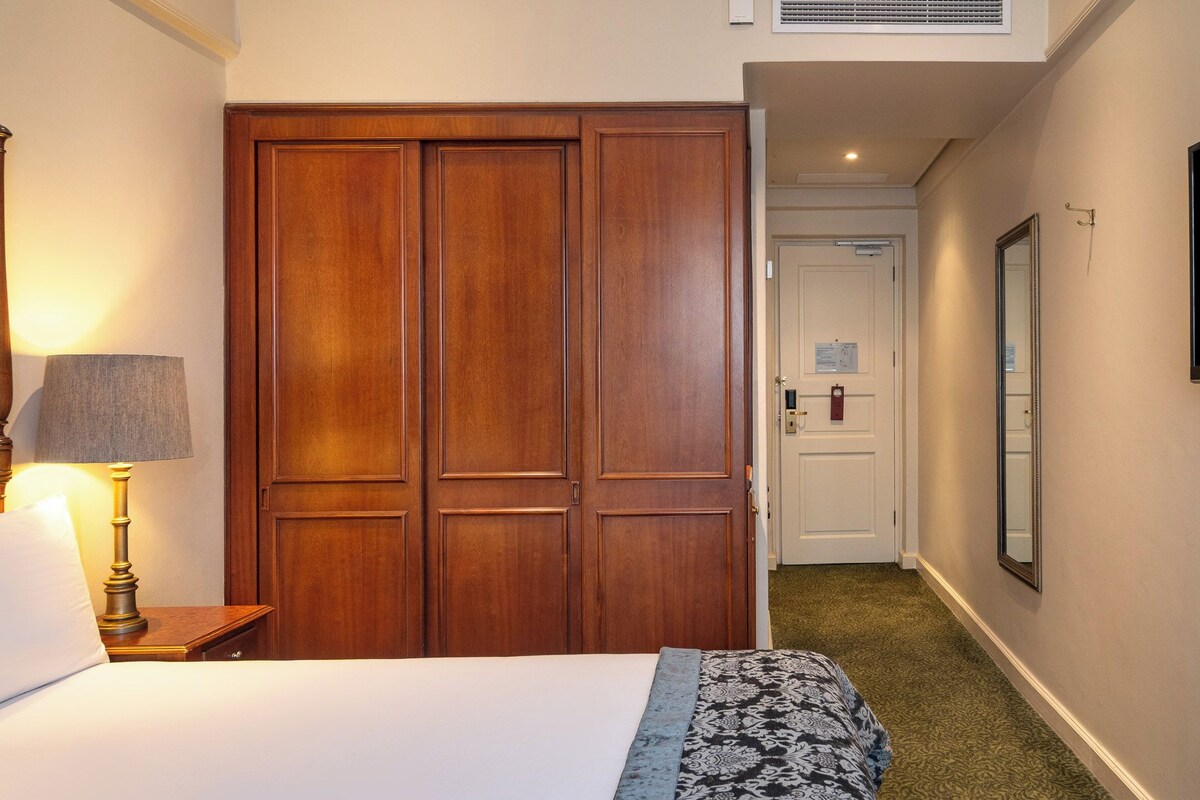 A hotel room with a wooden wardrobe, a bed, a bedside lamp, a television, and a carpeted hallway.
