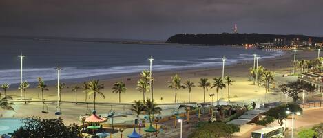 A beachside view with a road, palm trees, and a distant hill with a lighthouse.