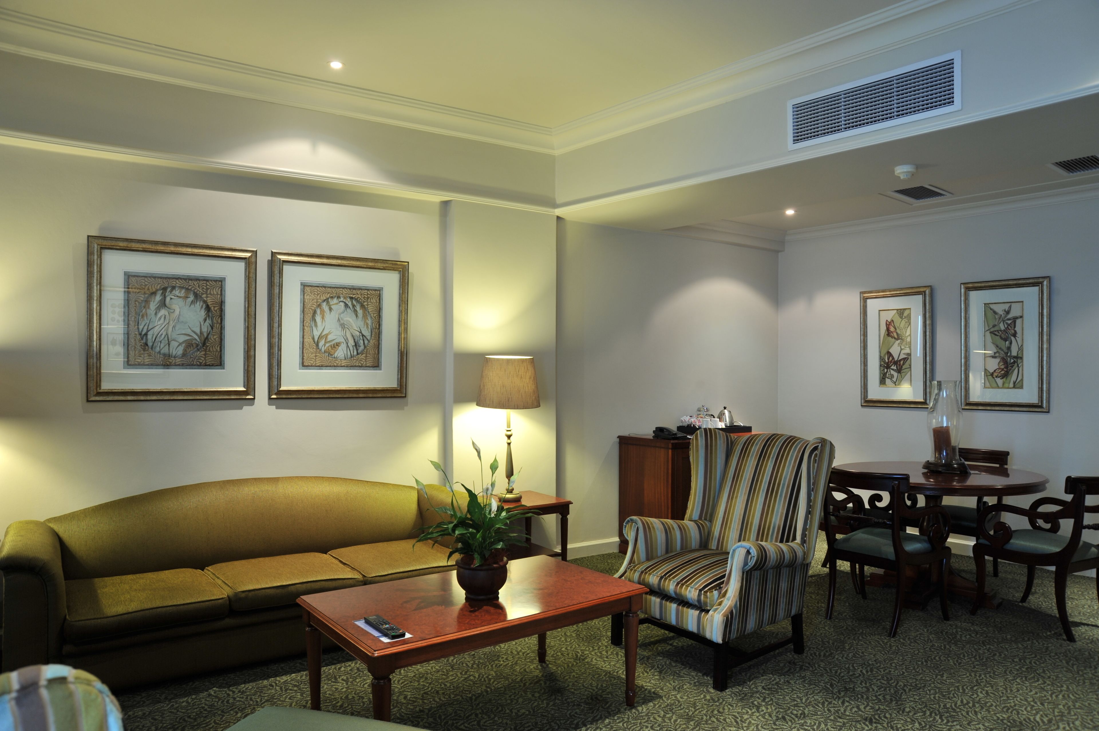 A living room with a sofa, armchair, coffee table, and dining table. There are framed pictures on the walls and a lamp on the side table.