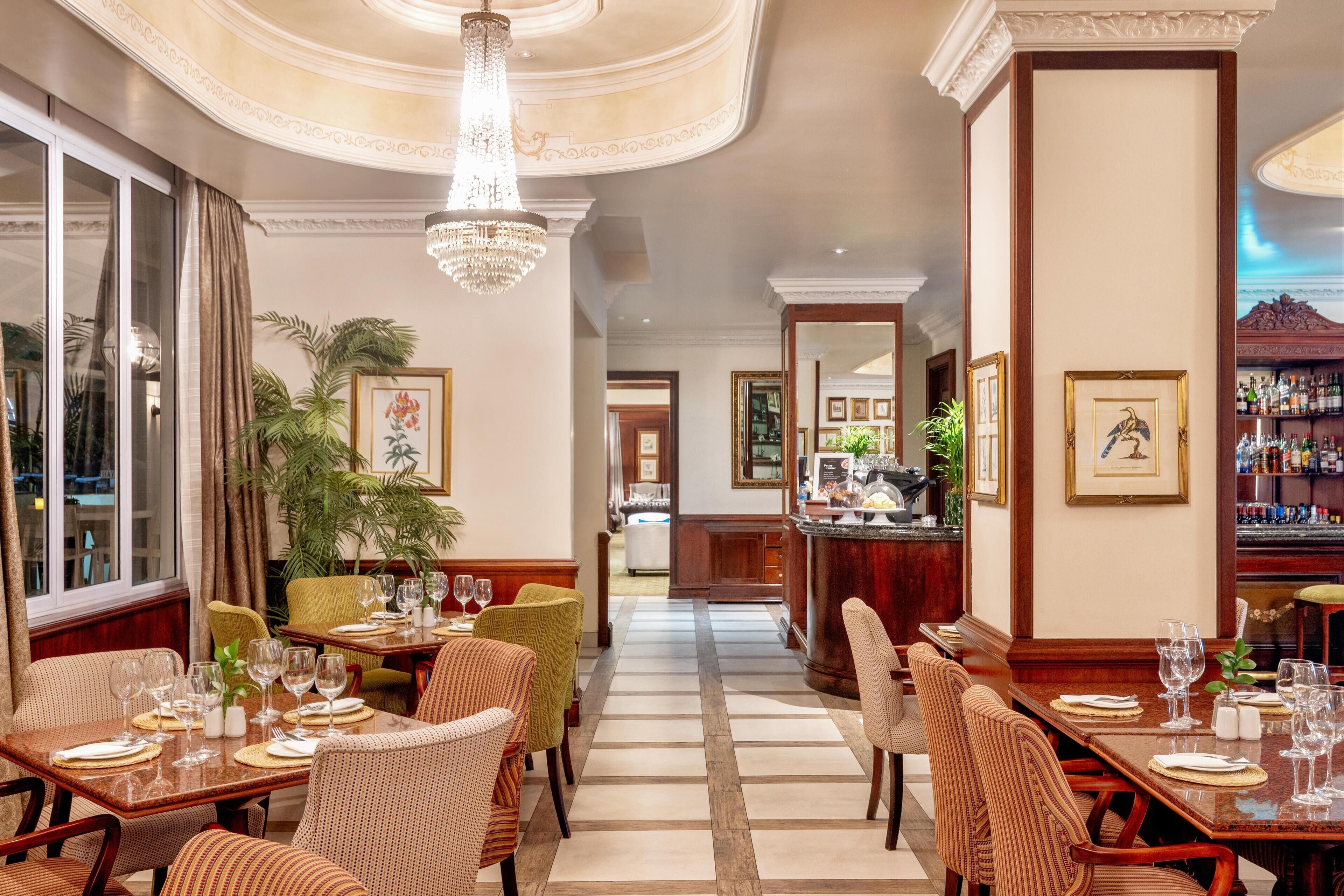 A hotel dining area with tables set for guests, a bar in the background, and a view of the pool through the window.