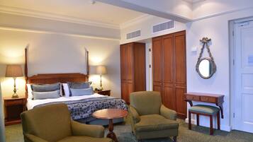 Una habitación de hotel con una cama, dos sillones, una mesita, una mesilla de noche, un espejo y un armario.