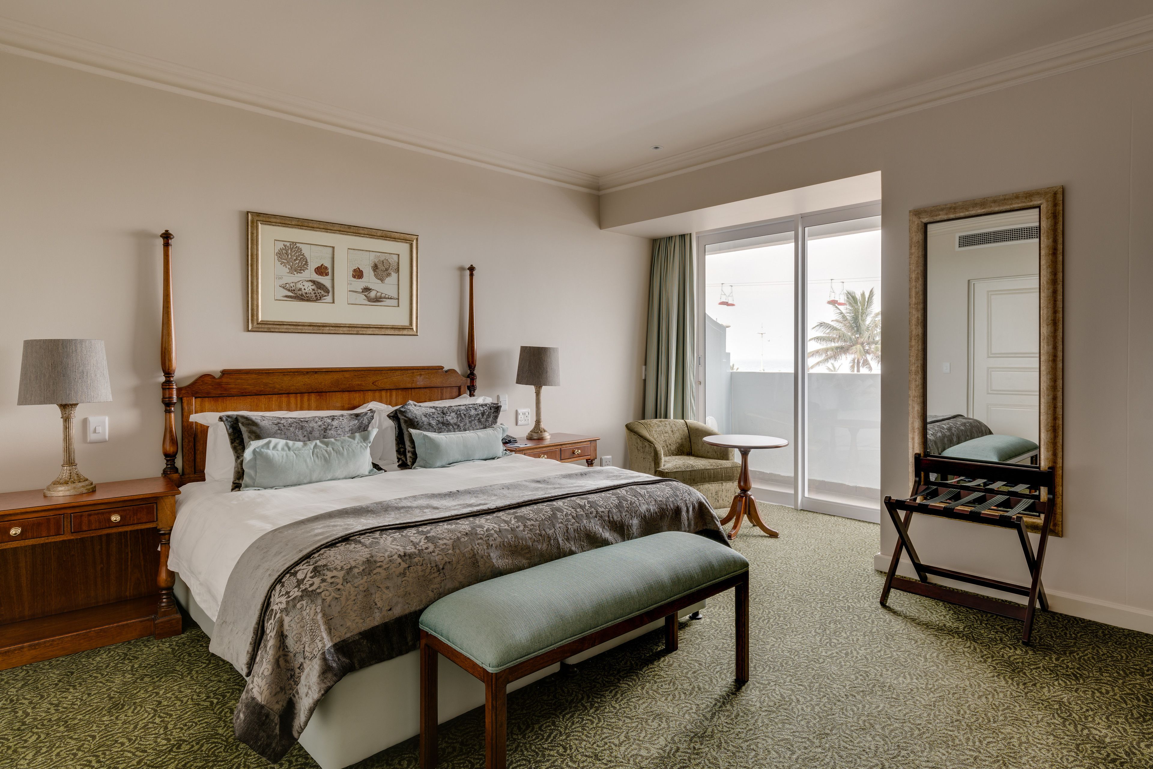 A hotel room with a large bed, a bench, a small table, a chair, and a mirror.