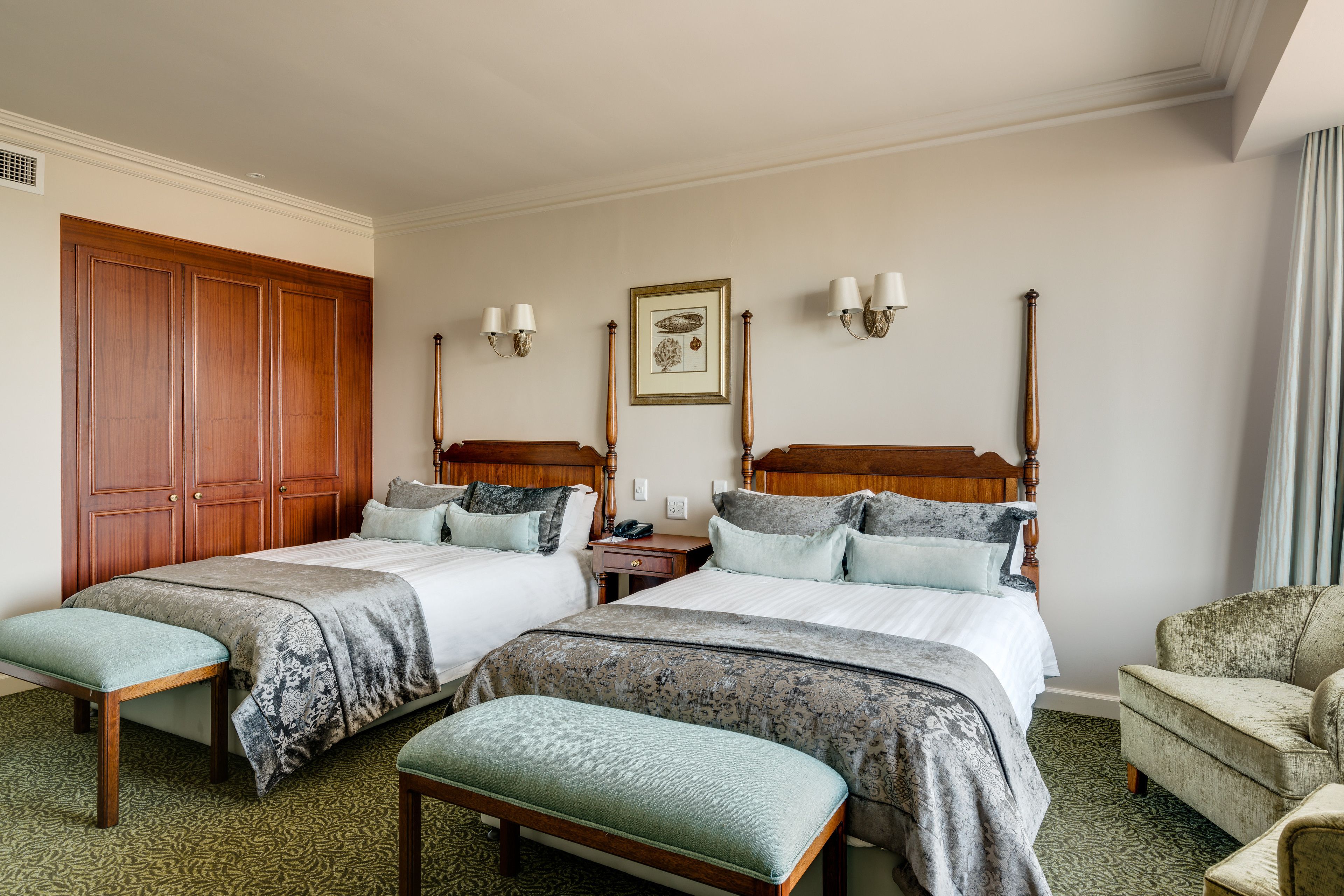 Habitación de hotel con dos camas, un armario de madera, un banco, una silla y un cuadro en la pared.
