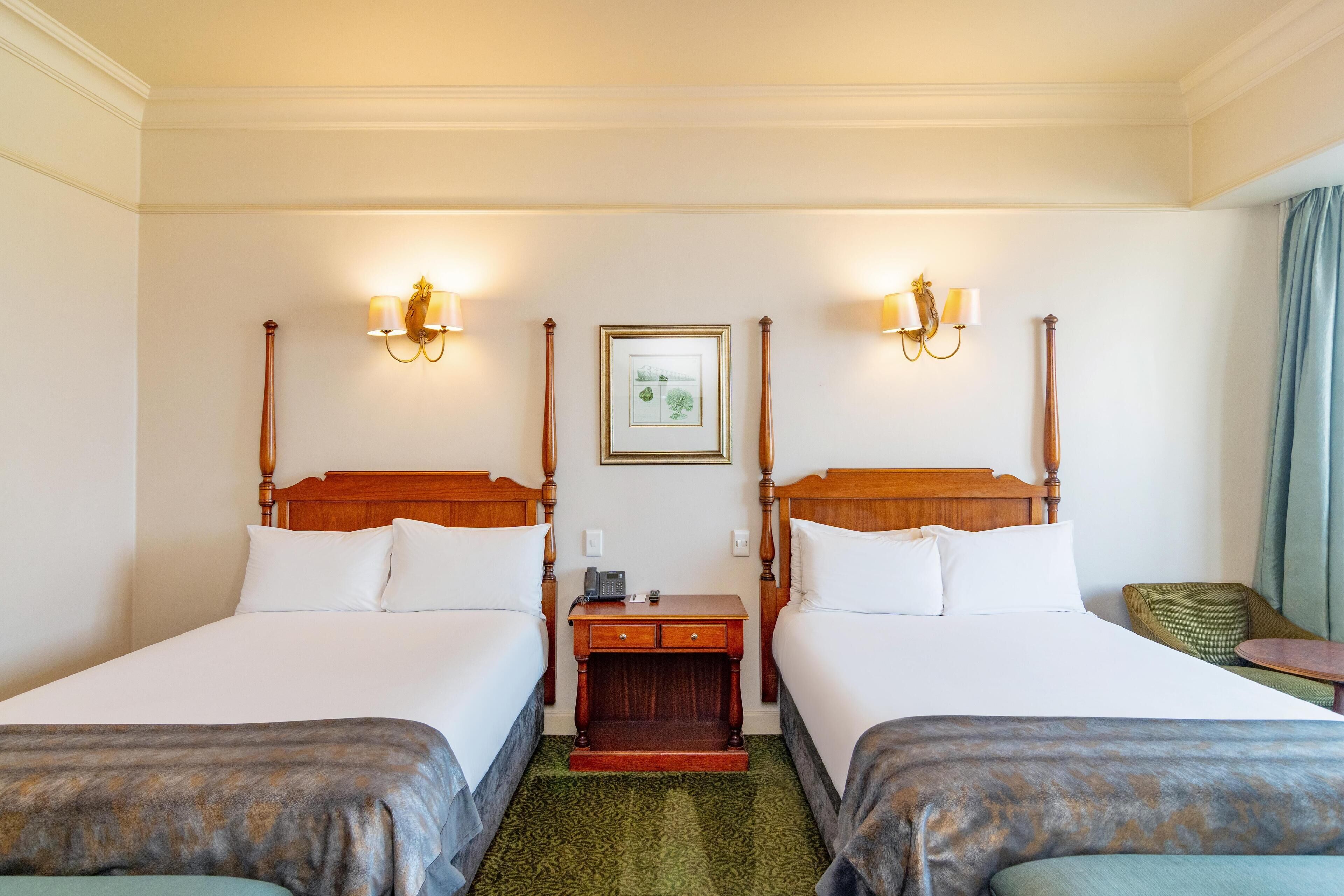 Une chambre d’hôtel avec deux lits simples, une table de chevet en bois, un tableau encadré au mur et une moquette verte.